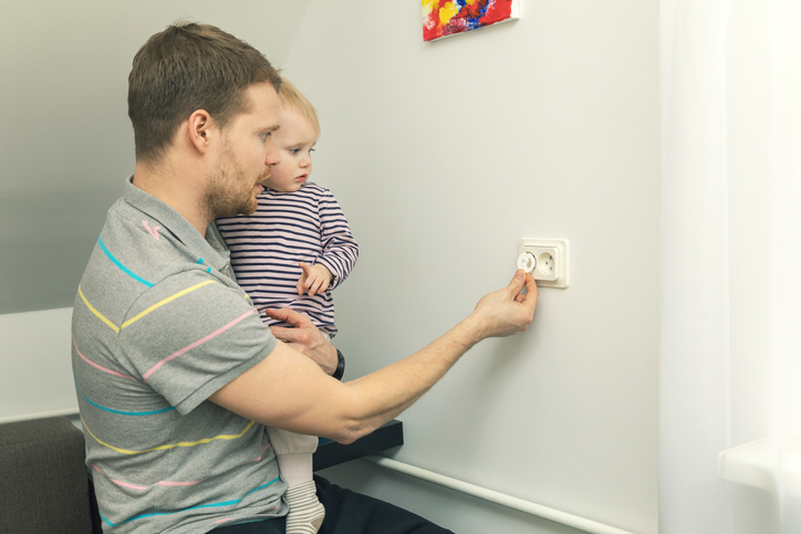 Tėtis laiko ant rankų mažą vaiką ir uždaro elektros kištuką.